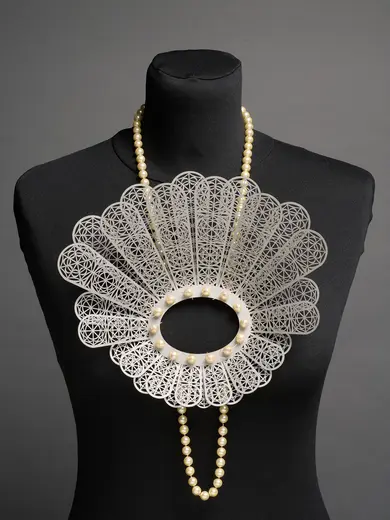 The image features a large, ornate necklace displayed on a black mannequin. The necklace has a lace-like fan shape with intricate patterns and is adorned with pearls, creating an elegant and striking appearance.