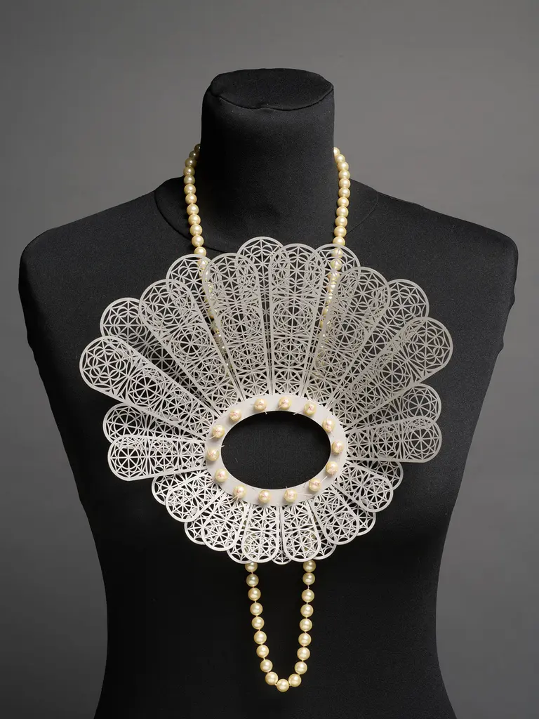 The image features a large, ornate necklace displayed on a black mannequin. The necklace has a lace-like fan shape with intricate patterns and is adorned with pearls, creating an elegant and striking appearance.