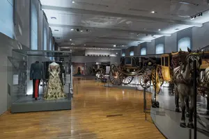 Eine Museumsausstellung mit historischen Kutschen und einer Vitrine, die eine elegante Robe und eine Uniform zeigt. Der Raum ist hell erleuchtet und hat einen holzgetäfelten Boden. Im Hintergrund sind weitere Exponate und eine Wand mit Kunstwerken sichtbar.