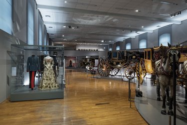 Eine Museumsausstellung mit historischen Kutschen und einer Vitrine, die eine elegante Robe und eine Uniform zeigt. Der Raum ist hell erleuchtet und hat einen holzgetäfelten Boden. Im Hintergrund sind weitere Exponate und eine Wand mit Kunstwerken sichtbar.