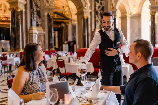 Ein Kellner serviert einem Paar am Tisch in einem eleganten Restaurant. Die Frauen trägt ein gestreiftes Oberteil, der Mann ein dunkles Hemd. Tischgedeck mit Gläsern und Servietten ist sichtbar. Eine stilvolle Atmosphäre mit opulenter Dekoration im Hintergrund.