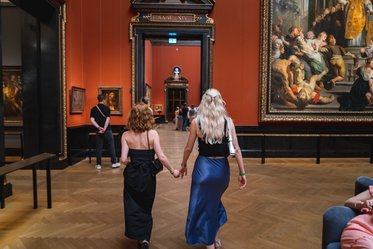 Zwei Frauen gehen Hand in Hand durch eine Kunstgalerie. Die eine trägt ein schwarzes Kleid, die andere einen langen blauen Rock und ein schwarzes Oberteil. Im Hintergrund sind mehrere Gemälde an den Wänden ausgestellt, und einige Besucher sind ebenfalls zu sehen.
