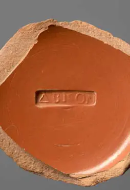 Ein runder, rotbrauner Tonteller mit einer eingravierten Inschrift in griechischen Buchstaben. Der Teller ist an einer Kante unregelmäßig abgebrochen, was auf eine historische Herkunft hinweist.