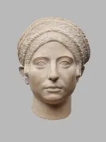 Eine antike Skulptur eines menschlichen Kopfes. Die Figur zeigt eine Frau mit geflochtenem Haar und einem neutralen Gesichtsausdruck. Sie hat große, runde Augen und einen klassischen, stilisierten Gesichtszug. Die Skulptur scheint aus hellem Stein gefertigt zu sein.