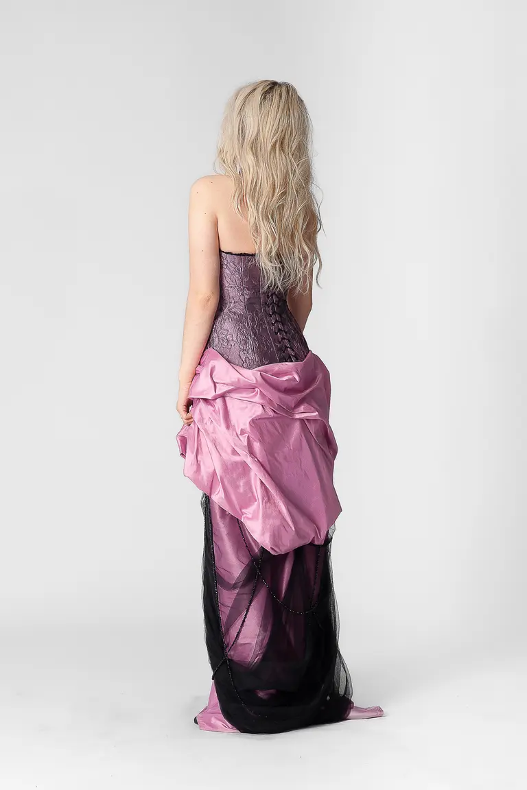 A model stands with her back to the camera, wearing a strapless gown. The dress features a fitted purple bodice and a flowing skirt in shades of pink and black. The background is white and minimalistic, emphasizing the gown's design.