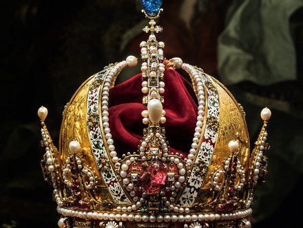 Eine kunstvoll gestaltete Krone aus Gold, verziert mit Perlen und Edelsteinen. Sie hat eine runde Form mit hohen, geschwungenen Spitzen. Im Inneren liegt rotes Samt, und auf der Spitze der Krone prangt ein blauer Edelstein. Die Krone steht auf einem roten Sockel.