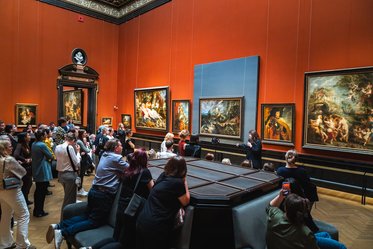 Ein Kunstbesucher führt eine Gruppe von Menschen durch einen museumartigen Raum. An den Wänden hängen mehrere Gemälde. Die Besucher sitzen und stehen in der Nähe einer zentralen Tischfläche, während sie den Erklärungen des Guides aufmerksam zuhören.
