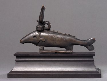 Eine bronzeartige Skulptur in Form eines Fisches mit einem oberen Handgriff. Die Figur steht auf einer rechteckigen Basis und zeigt detaillierte Merkmale wie die Kiemen und eine gezackte Rückenlinie.