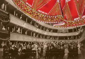 Eine historische Theateraufführung mit einem vollem Zuschauerraum. Elegant gekleidete Personen sitzen auf verschiedenen Ebenen, während ein beeindruckendes rotes und goldverziertes Bühnenlicht im Fokus steht. Die Atmosphäre wirkt festlich und feierlich.