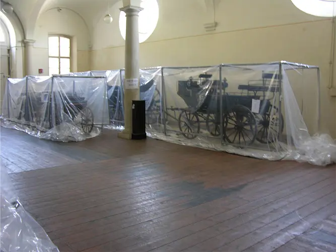 Im Bild sind mehrere historische Kutschen in einem Raum zu sehen, die durch durchsichtige Plastikfolie geschützt sind. Sie stehen auf einem Holzboden und sind hinter transparenten Abdeckungen sichtbar. Der Raum hat hohe Decken und große Fenster.