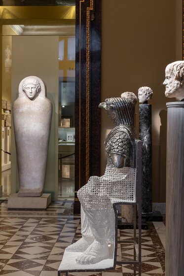 Eine Ausstellung im Museum zeigt verschiedene Skulpturen: Im Vordergrund sitzt eine Statue mit römischer Rüstung, während im Hintergrund eine große menschliche Figur und mehrere Kopfskulpturen aus Stein stehen. Der Boden ist mit einem dekorativen Mosaikmuster gestaltet.