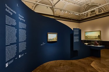Ein Ausstellungsraum mit blauen Wänden, die Texte über venezianische Kunst zeigen. An der Wand hängen zwei Gemälde, eines davon ist deutlich sichtbar. Der Boden ist aus Holz, und es gibt eine Vitrine, die Kunstobjekte präsentiert.