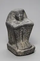 Schwarzer Stein mit der Darstellung eines Kopfes, der eine ägyptische Figur darstellt. Der Körper hat Hieroglyphen, die an den Seiten eingraviert sind. Die Figur hat lange, geflochtene Haare und einen eindrucksvollen Gesichtsausdruck.