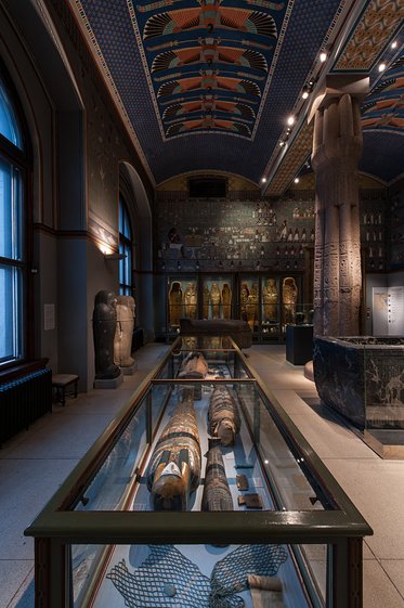 Ein Raum in einem Museum mit antiken ägyptischen Artefakten. In Vitrinen sind Sarkophage und andere Exponate ausgestellt. Die Wände sind mit farbenfrohen Wandmalereien verziert, und eine hohe Decke trägt zum historischen Ambiente bei.