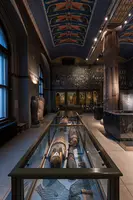 Ein Raum in einem Museum mit antiken ägyptischen Artefakten. In Vitrinen sind Sarkophage und andere Exponate ausgestellt. Die Wände sind mit farbenfrohen Wandmalereien verziert, und eine hohe Decke trägt zum historischen Ambiente bei.