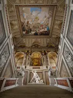 Ein imposanter Treppenaufgang in einem prunkvollen Gebäude. Die Wände sind reich verziert, und an der Decke befindet sich ein beeindruckendes Gemälde. Im Vordergrund steht eine massive Skulptur, die eine Szene mit Figuren darstellt, umgeben von kunstvollen Details.