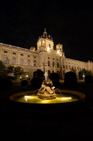 Eine beleuchtete Brunnenanlage steht im Vordergrund, während im Hintergrund ein beeindruckendes Gebäude bei Nacht zu sehen ist. Die Fassade des Gebäudes ist detailliert und elegant gestaltet, umgeben von Bäumen und Sträuchern.