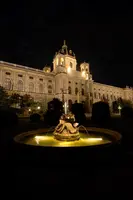 Eine beleuchtete Brunnenanlage steht im Vordergrund, während im Hintergrund ein beeindruckendes Gebäude bei Nacht zu sehen ist. Die Fassade des Gebäudes ist detailliert und elegant gestaltet, umgeben von Bäumen und Sträuchern.