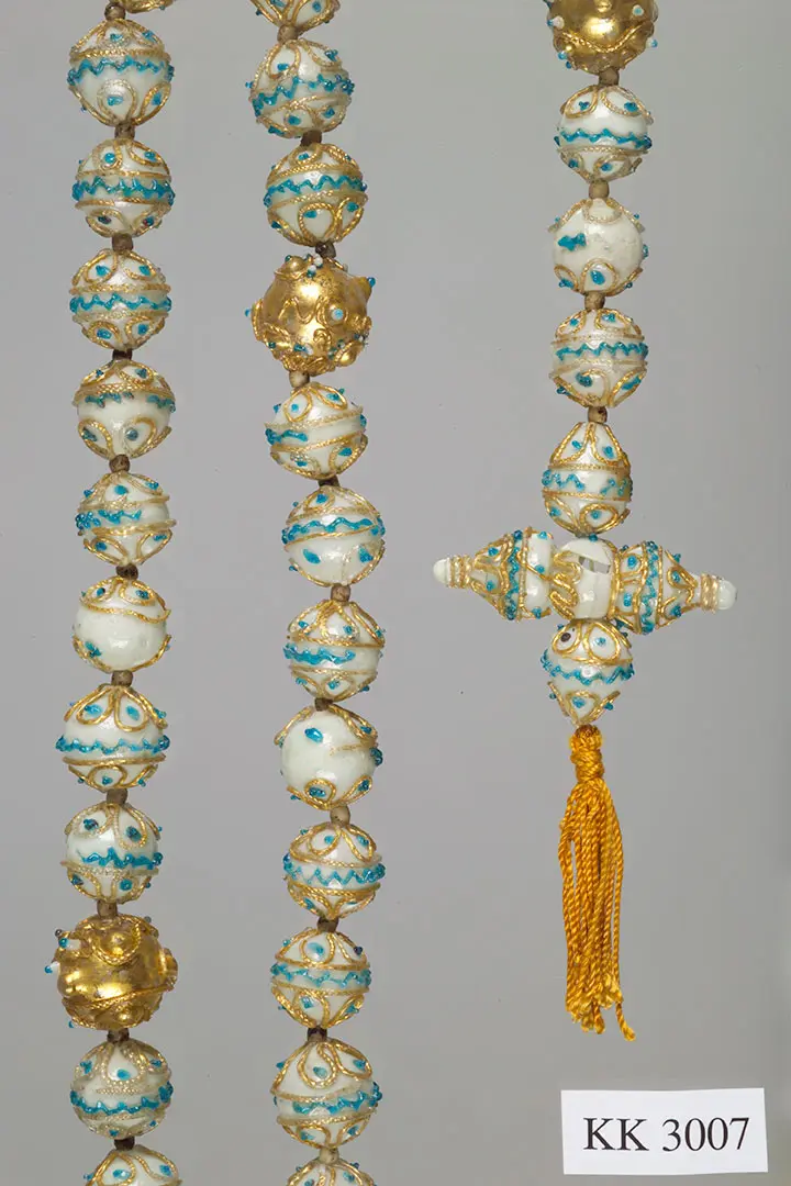 Das Bild zeigt eine dekorative Kette mit verschiedenen Perlen in Gold und Weiß, verziert mit blauen Akzenten. In der Mitte befindet sich ein auffälliger Anhänger mit ähnlichen Designelementen. Die Kette hat eine elegante und kunstvolle Ausstrahlung.