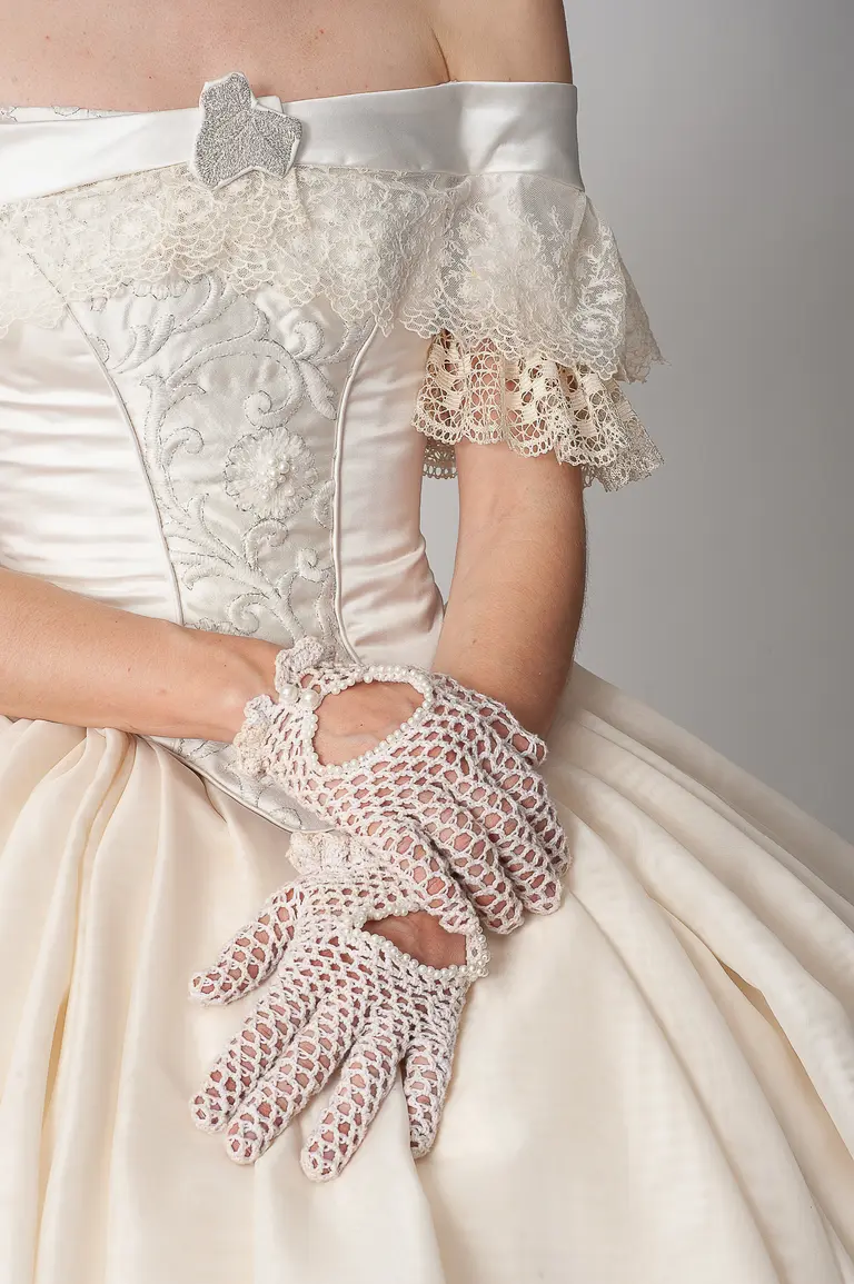 A close-up of a person's arms wearing a vintage-style gown with lace detailing and puffed sleeves. The hands are adorned with delicate, lace gloves, resting elegantly on fabric of a full, flowing skirt. The overall look embodies a classic and formal aesthetic.