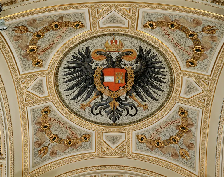 Das Bild zeigt eine kunstvoll gestaltete Decke mit einem Doppeladler-Wappen in der Mitte. Das Wappen ist rot mit einem weißen Feld, umgeben von detaillierten floralem Design und goldenen Elementen. Es vermittelt ein Gefühl von historischer Pracht und Symbolik.