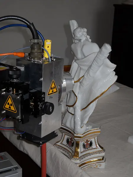 Ein strahlender Scanner steht neben einer weißen Porzellanstatue, die eine weibliche Figur in eleganter Pose darstellt. Die Statue ist auf einem kunstvoll gestalteten Sockel platziert. Der Scanner hat Warnschilder, die auf die Verwendung von Strahlung hinweisen.