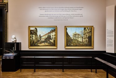 Zwei große Gemälde hängen an einer Wand in einem Museum. Links ist eine Stadtansicht mit Gebäuden und Menschen. Rechts zeigt das andere Gemälde ein prächtiges Gebäude. Darunter stehen Informationen über die Kunstwerke. Im Vordergrund sieht man eine Vitrine mit einem weißen Bust.