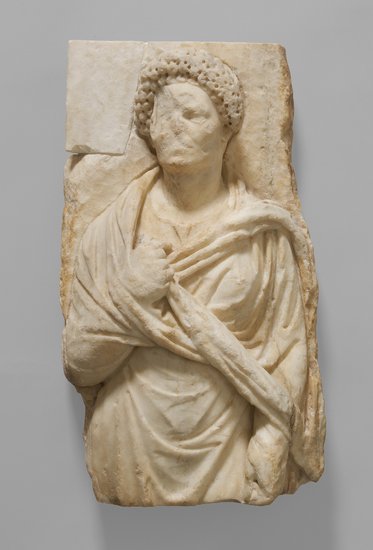 Eine antike marmorne Skulptur zeigt eine figurative Darstellung einer Person in frontalem Stand. Die Figur trägt eine Draperie und hat eine zur Brust gefaltete Hand. Der Kopf ist teilweise beschädigt, die Details sind jedoch klar erkennbar.