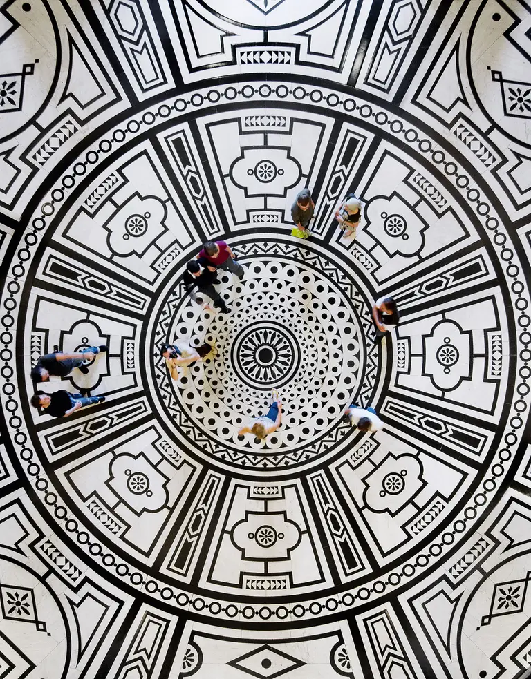 Ein Rundmuster in Schwarz-Weiß auf dem Boden im KHM ist zu sehen. Mehrere Personen stehen und bewegen sich innerhalb dieses Musters. Das Design ist geometrisch und symmetrisch, mit auffälligen Kreisen und floralen Motiven im Mittelpunkt.