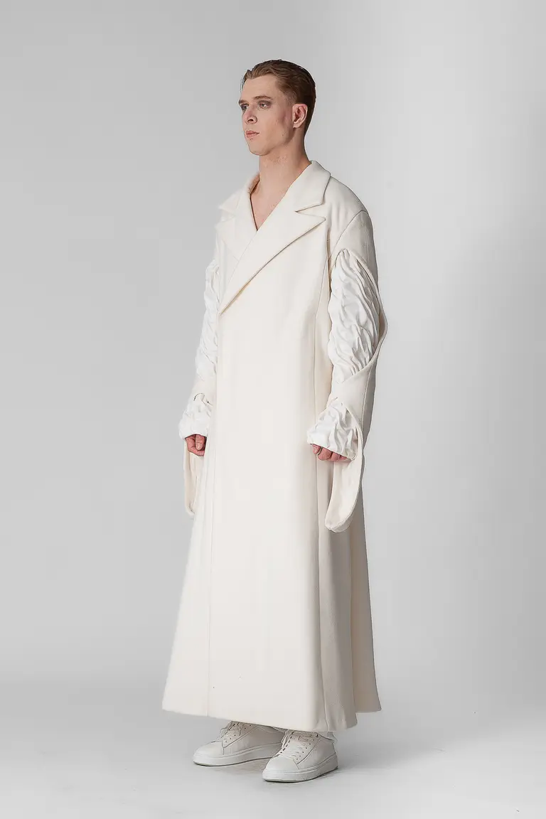 A model stands in profile wearing a long, oversized white coat with wide sleeves and a double-breasted design. The sleeves feature intricate detailing, contrasting with the simple, elegant style of the coat. The model pairs the outfit with white sneakers, set against a neutral background.