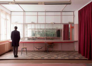 Eine Person steht in einem leeren Raum mit roten Vorhängen und einem Spiegel. Der Raum hat eine schlichte Einrichtung mit einem Tisch und einem Stuhl. Das Licht fällt durch große Fenster und beleuchtet den minimalistischen Raum.