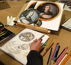 A hand holds a pencil over a sketchbook with drawn portraits. Nearby, there are reference images of classic paintings. Colored pencils and art supplies are arranged around the workspace, indicating an artistic environment focused on sketching and studying portraits.