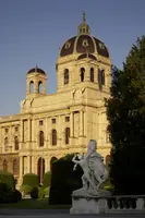 Das Bild zeigt ein historisches Gebäude mit einer markanten Kuppel und ornamentierten Fassaden. Im Vordergrund steht eine Statue, die einen Reiter darstellt. Die Szene ist von grünen Bäumen und Sträuchern umgeben, und das warme Licht hebt die Architektur hervor.
