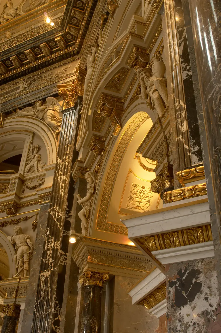Das Bild zeigt kunstvoll gestaltete Wände mit reich verzierten Säulen und Statuen. Goldene Verzierungen und prächtige Details sind deutlich sichtbar, die eine opulente Architektur darstellen. Licht strahlt von oben und hebt die kunstvollen Elemente hervor.