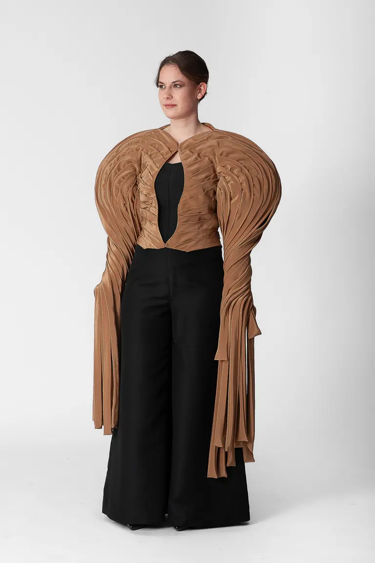 A model stands against a plain white background, wearing a striking fashion outfit. The ensemble features an elaborate brown top with large, textured sleeves and a fitted black pant. The design emphasizes unique shapes and a dramatic silhouette.