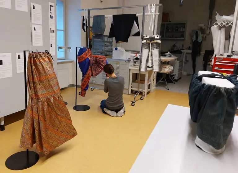 A person is crouching on the floor in a fashion design studio, examining a colorful garment displayed on a rack. Two other garments, a patterned skirt and a pair of denim pants, are visible on stands nearby, along with various sewing materials in the background.