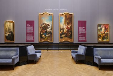Ausstellung mit Gemälden von Giovanni Battista Tiepolo. In der Mitte hängen zwei große Gemälde in goldenen Rahmen, flankiert von Leinwandbeschreibungen. Links steht ein weiteres Gemälde, rechts ein weiteres, während im Vordergrund blau gepolsterte Sitze sichtbar sind.