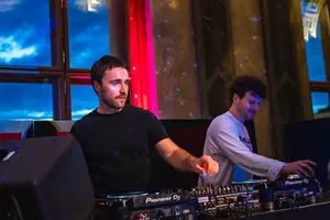 Zwei DJs stehen hinter einem Mischpult in einem beleuchteten Raum. Der DJ links trägt ein schwarzes T-Shirt und konzentriert sich auf die Musik, während der DJ rechts in einem weißen Sweatshirt lächelt. Der Hintergrund zeigt eine stimmungsvolle Beleuchtung und einen hellen Himmel.