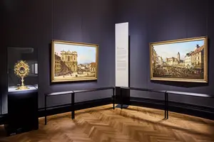 Ein Ausstellungsraum mit zwei Gemälden an der Wand, die städtische Szenen zeigen. In der Mitte steht ein Glaskasten mit einem kunstvollen, goldenen Monstranz. Dazwischen befindet sich ein informationstafel auf einem vertikalen Ständer. Der Boden ist aus parkett.