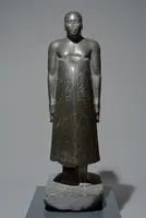 Eine schwarze Statue eines stehenden Mannes mit glattem Körper und langen Armen. Er trägt ein schlichtes, langes Gewand und hat einen ernsthaften Gesichtsausdruck. Auf dem Gewand sind Hieroglyphen zu sehen. Die Statue ist auf einem Sockel platziert und hebt sich vor einem neutralen Hintergrund ab.