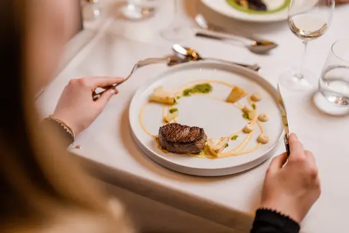 Auf dem Bild sieht man einen gedeckten Tisch mit einem Gourmetgericht auf einem Teller. Eine Person hält Besteck in der Hand, während sie das Fleischgericht mit Beilagen betrachtet. Der Teller ist kunstvoll angerichtet, umgeben von Gläsern und Geschirr.