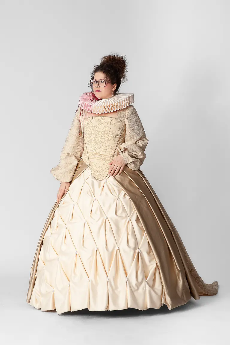 A person wearing an elaborate historical-inspired gown with a voluminous skirt and intricate detailing. The dress is cream and gold, complemented by a ruffled collar in soft pink tones. They have curly hair and glasses, standing against a plain background.