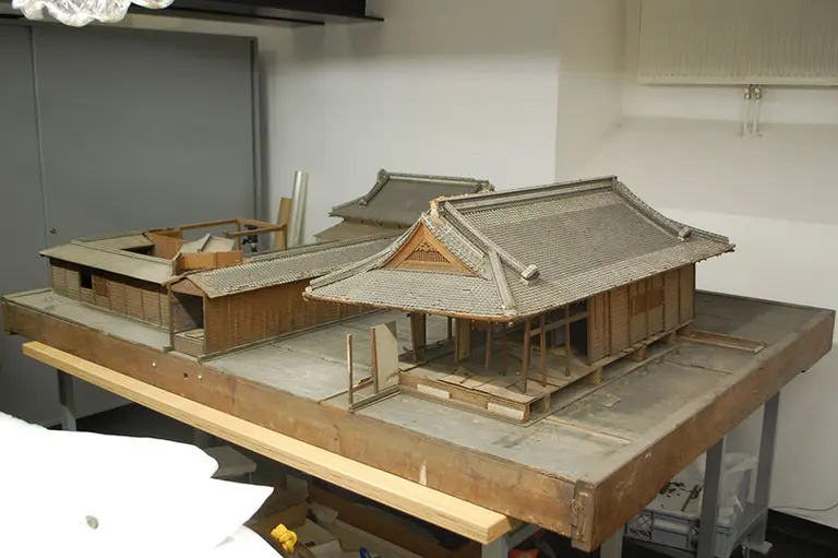 Das Bild zeigt ein detailliertes Modell eines traditionellen japanischen Hauses mit zwei Gebäuden auf einer Holzplatte. Die Dächer sind mit Ziegeln gedeckt, und die Wände sind aus Holz. Der Hintergrund zeigt einen neutralen Raum mit wenig Ablenkung.