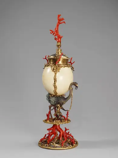 Ein kunstvolles Gefäß mit einer ovalen, cremefarbenen Form, umgeben von goldenen Verzierungen. Darunter befindet sich eine Skulptur mit rotem Korallenartigem Element und einem fantastischen, geschwungenen Fuß. Das Gefäß hat zwei Deckel und ist reich dekoriert.