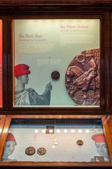 Ein Ausstellungsdisplay zum Thema Kopfbedeckungen in der Renaissance. Links ist ein Bild eines Mannes mit rotem Hut, daneben ein Relief einer Reiterfigur. Unten befinden sich verschiedene Münzen und Medaillons, die die Bedeutung von Hüten in der Mode hervorheben.