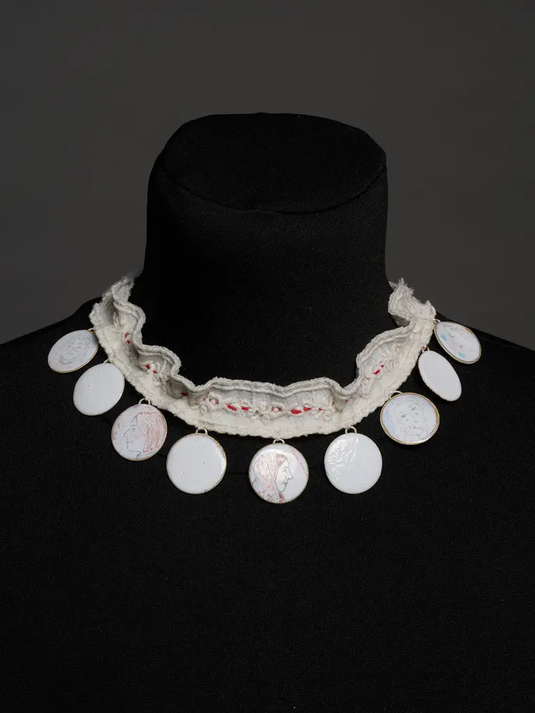 A close-up of a decorative necklace displayed on a mannequin. The necklace features a ruffled fabric base with several round, flat pendants hanging from it, some with intricate designs and others plain. The background is dark, highlighting the necklace's details.