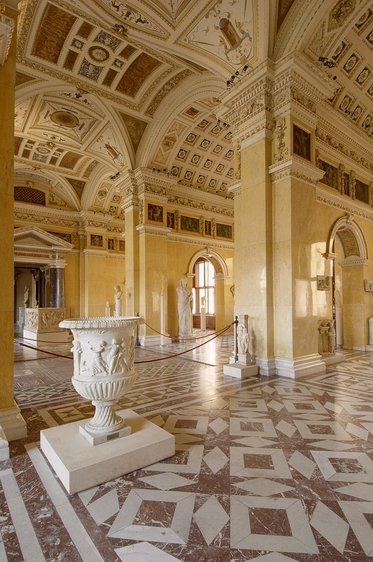 Eine weite Innenansicht eines eleganten Raumes mit hohen Decken und aufwendigen Wandmalereien. In der Mitte steht ein verziertes, antikes Podest. Umgeben von Skulpturen und Säulen, strahlt der Raum historischen Charme aus. Der Boden ist mit einem geometrischen Muster gestaltet.