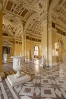 Eine weite Innenansicht eines eleganten Raumes mit hohen Decken und aufwendigen Wandmalereien. In der Mitte steht ein verziertes, antikes Podest. Umgeben von Skulpturen und Säulen, strahlt der Raum historischen Charme aus. Der Boden ist mit einem geometrischen Muster gestaltet.