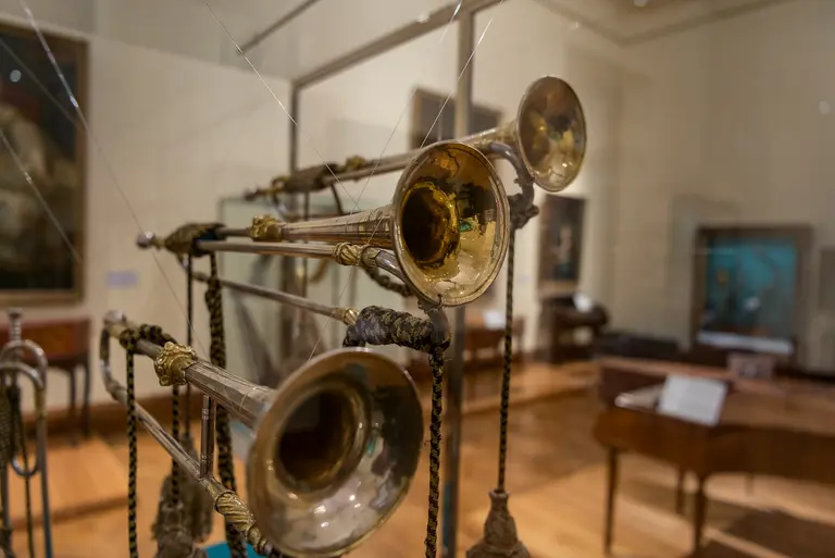 A display of vintage brass trumpets hanging from strings in a museum. The polished instruments shine under the light, showcasing their intricate details and craftsmanship against a neutral background of a gallery. The atmosphere suggests a celebration of musical history.