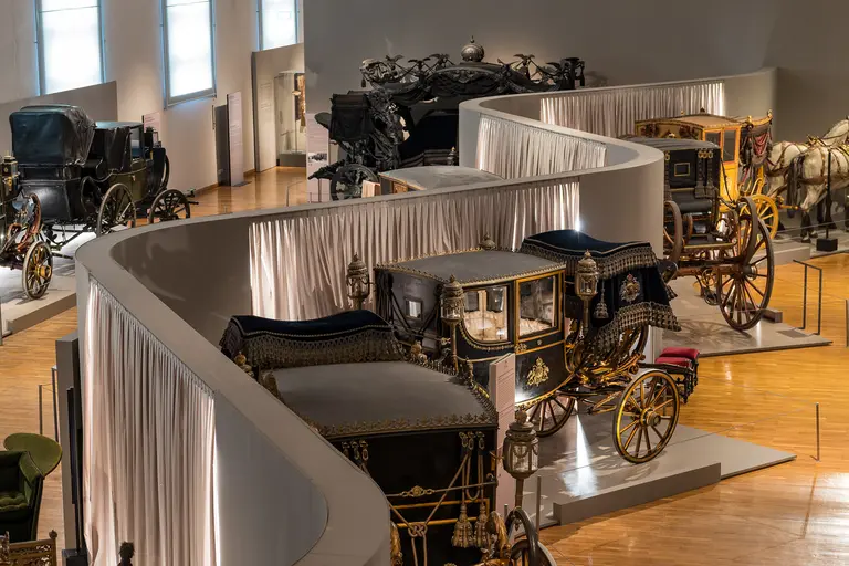 An exhibit featuring various historical carriages in a museum setting. The carriages are displayed on a wooden floor, arranged behind a curved partition draped with fabric. Natural light illuminates the surroundings, highlighting the intricate designs of the vehicles.